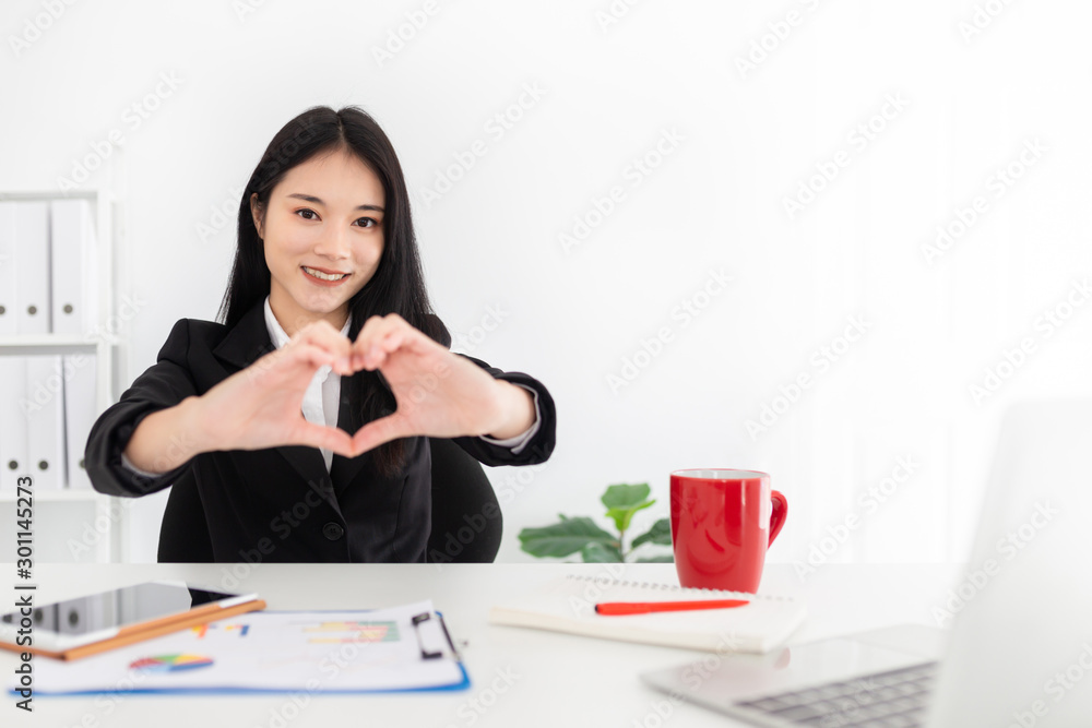 有魅力的亚洲官员用手展示心形标志，她坐在办公室里，她用笔记本电脑和th
