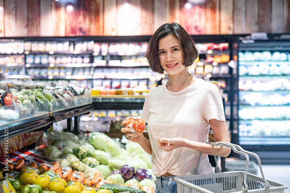 年轻的亚洲美女拿着购物篮走在超市里。她手里拿着番茄。