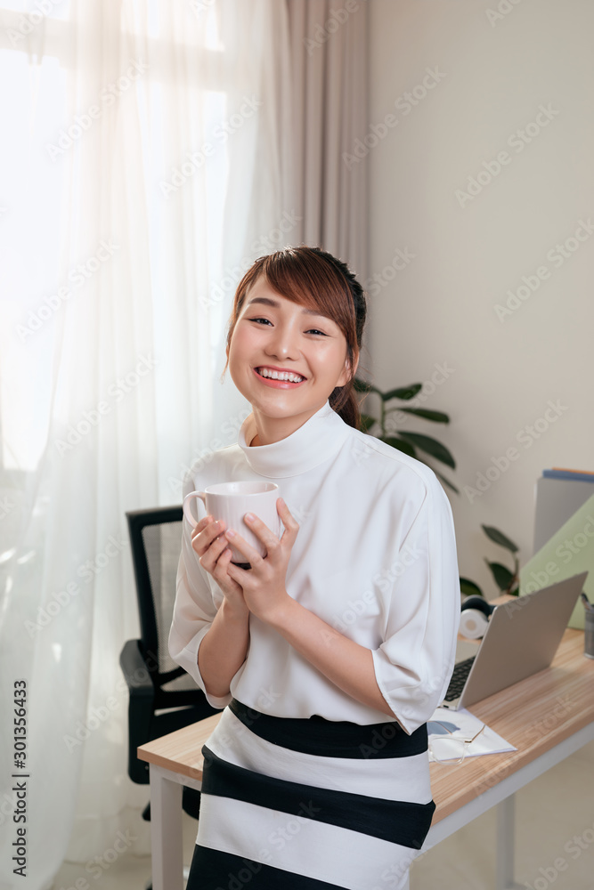 年轻的亚洲女商人坐在办公桌上，手里拿着咖啡杯