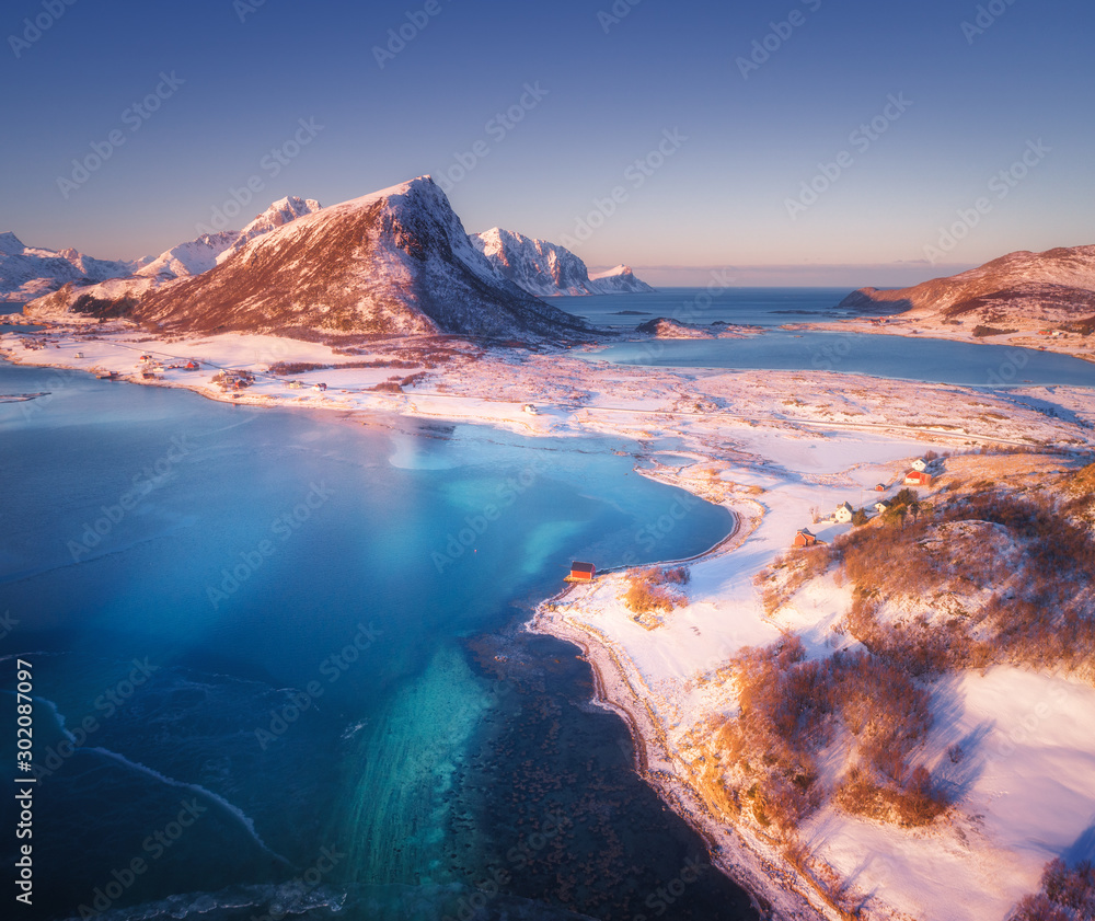 日落时白雪皑皑的山脉、房屋、碧水和蓝天的鸟瞰图。冬季景观