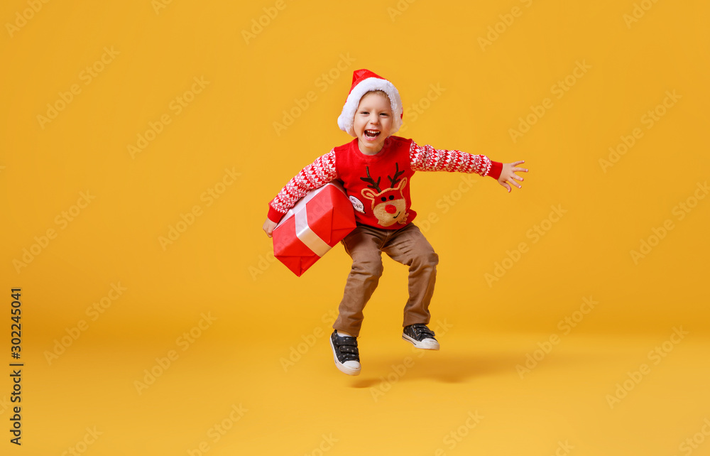 happy funny child boy in red Christmas reindeer costume jumping  with gift on yellow   background.