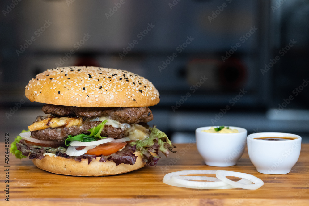 A big fresh colorful burger with meat, egg, salad, onion rings and dips on a wooden tray