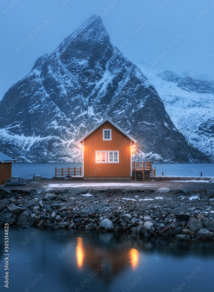 Red rorbu on sea coast and snow covered mountain at night. Lofoten islands, Norway. Moody winter lan