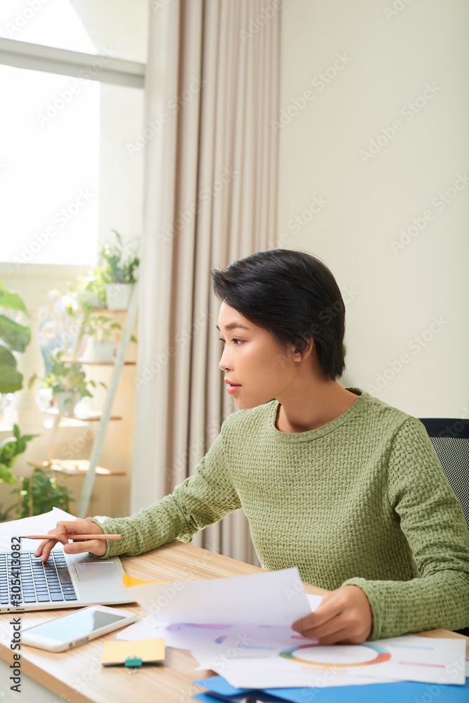 年轻的亚洲女性坐在办公桌上，看着笔记本电脑屏幕