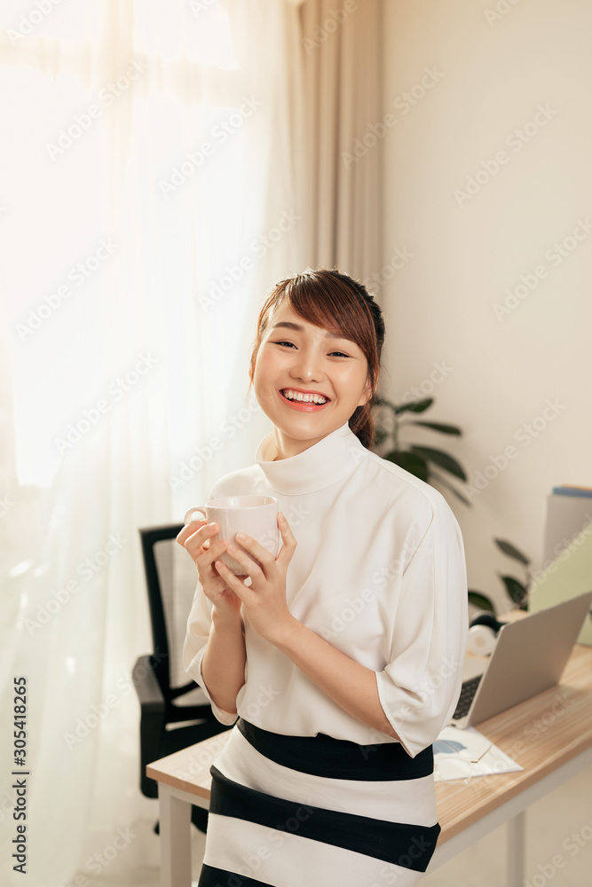 年轻的亚洲女商人坐在办公桌上，手里拿着咖啡杯