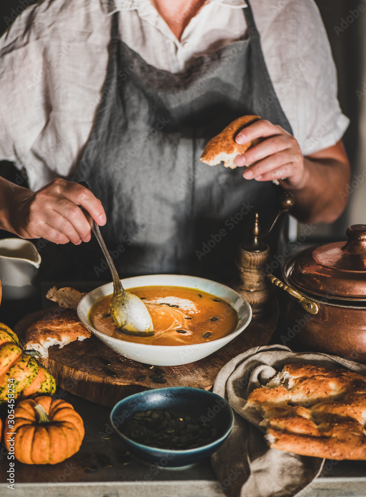 Eldery woman in grey linen apron mixing cream with Autumn seasonal pumpkin cream soup with spoon in 