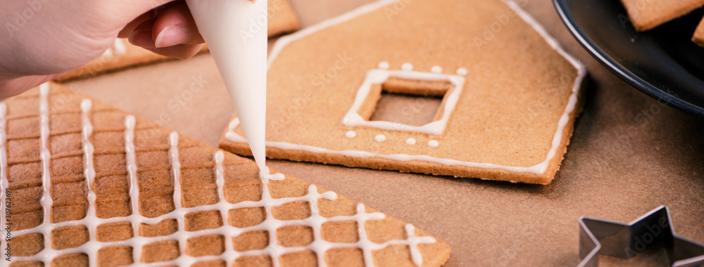 Woman is decorating gingerbread cookies house with white frosting icing cream topping on wooden tabl