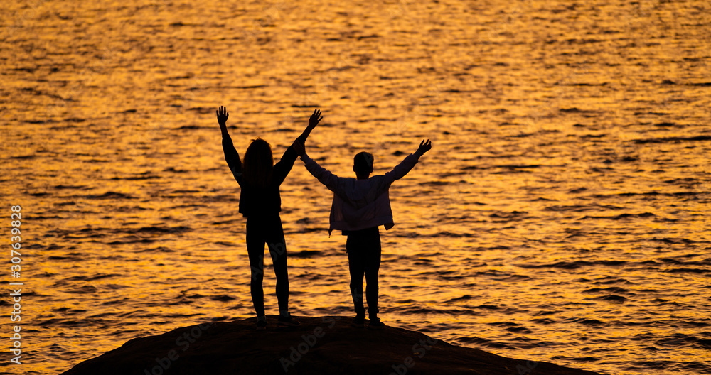 两个年轻人站在水面前的岩石上，举起双手看着日落。再次欢呼。