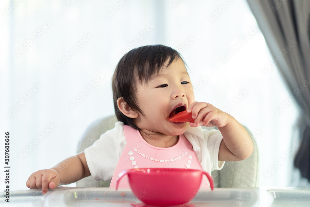 坐在婴儿高脚椅上喂奶的小可爱，在家里面带微笑地抱着西瓜。孩子