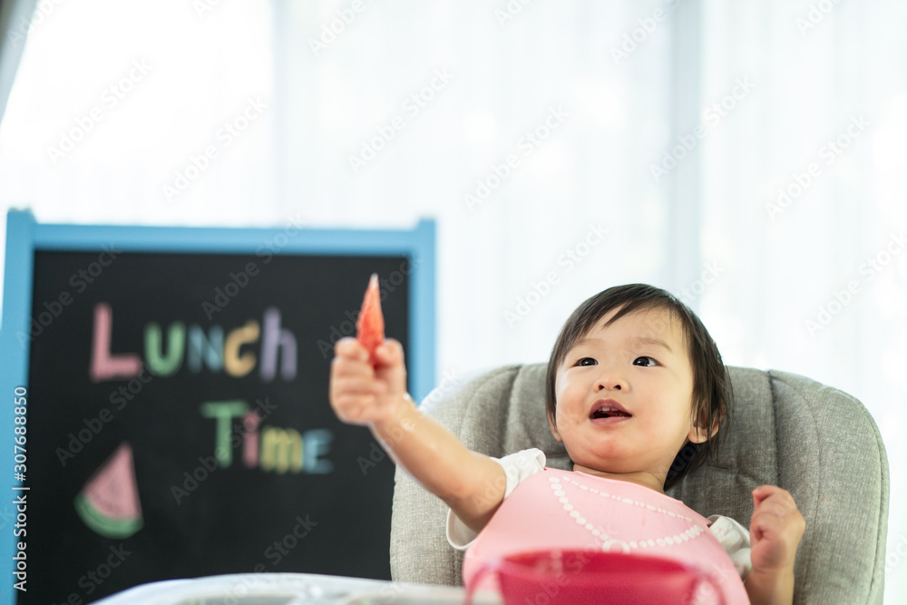 坐在婴儿高脚椅上的小可爱，在家里面带微笑地抱着西瓜。孩子