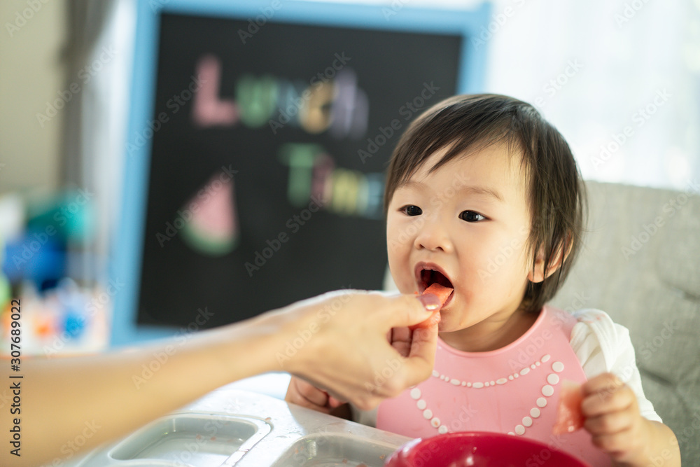 Mother feeding watermelon to young cute kid on baby high chair feeding seat at home. The child enjoy