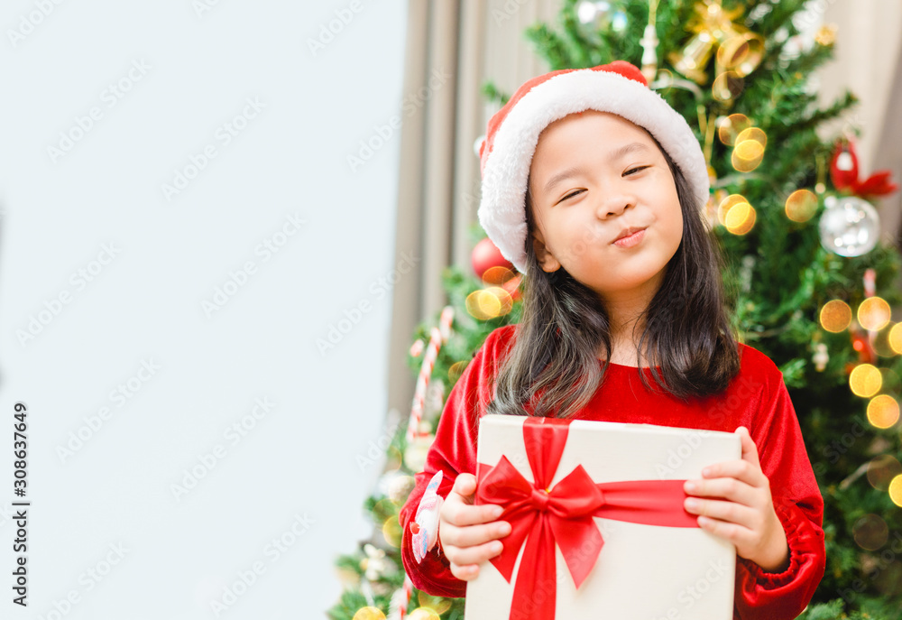 亚洲小女孩微笑着，兴奋地拿着圣诞树上的红色礼盒，白色背景。