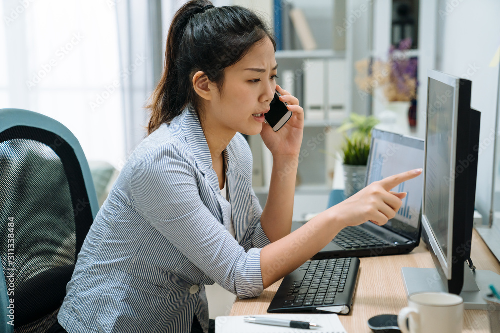皱着眉头的亚裔日本女员工在办公室用笔记本电脑工作，一边打手机