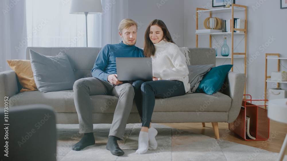 可爱的年轻情侣坐在舒适公寓的沙发上使用笔记本电脑。情侣冲浪