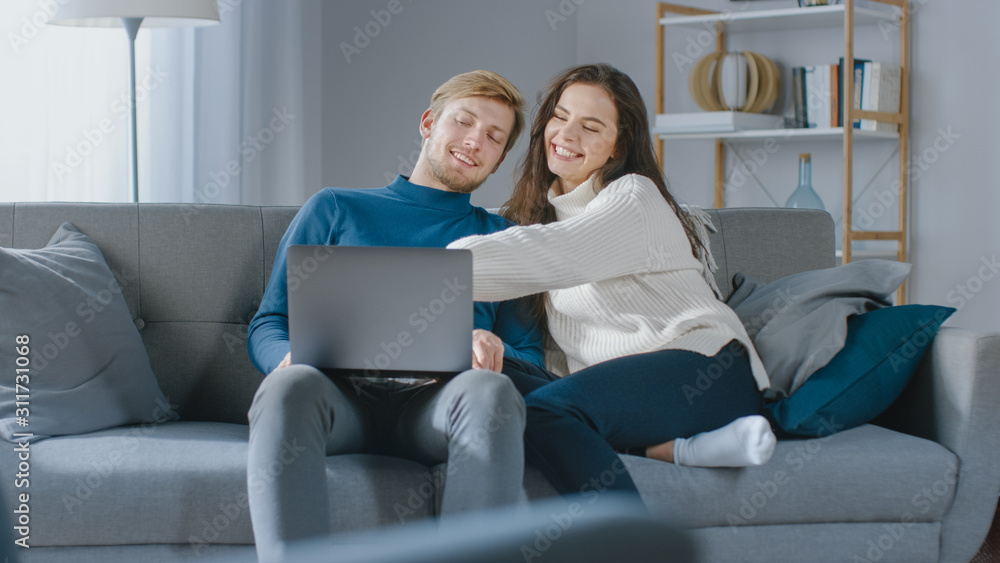 可爱的年轻夫妇在舒适的公寓里坐在沙发上使用笔记本电脑，玩得开心，大笑