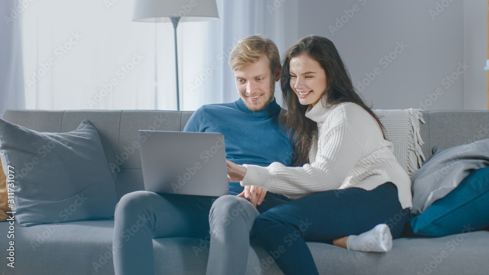 Cute Young Couple Use Laptop Computer, Have Fun, Laughs, while Sitting on the Couch in the Cozy Apar