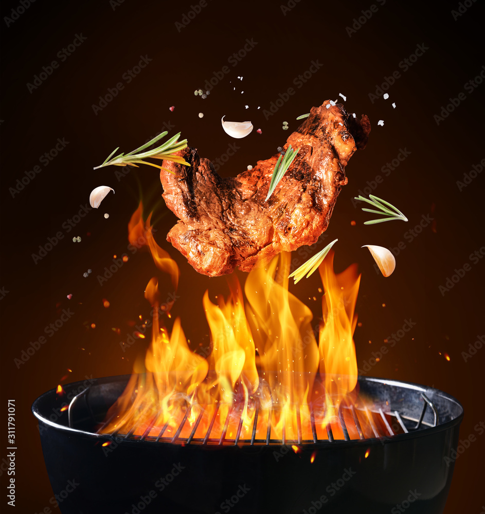 在黑暗背景下，带着火焰的烧烤架上掉落美味的肉和香料