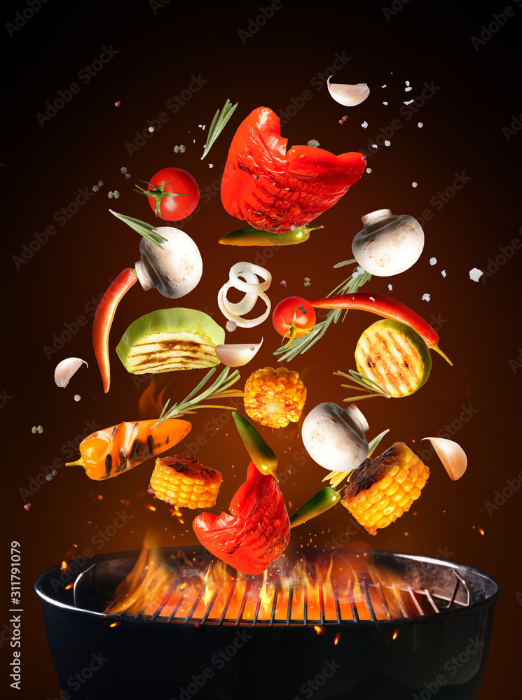 美味的蔬菜和香料掉落在烧烤架上，火焰背景为深色