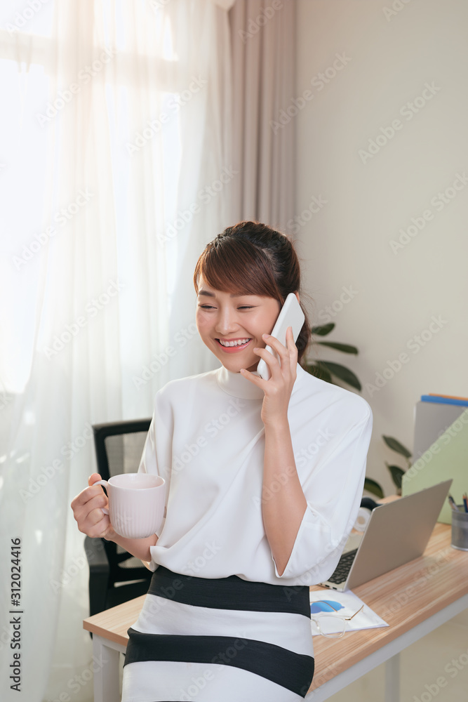 年轻的亚洲女商人在办公室的桌子边喝咖啡边打电话