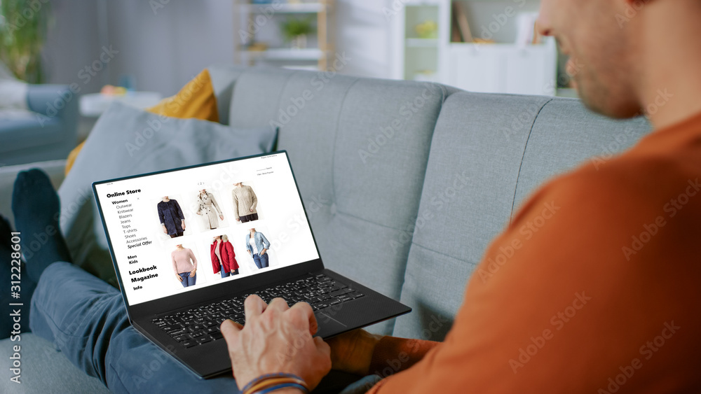 英俊的年轻人坐在家里使用笔记本电脑，浏览网上零售商的服装销售存储库