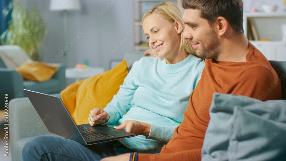 可爱的年轻夫妇坐在家里的沙发上，他们用笔记本电脑玩得很开心。美丽的肖像