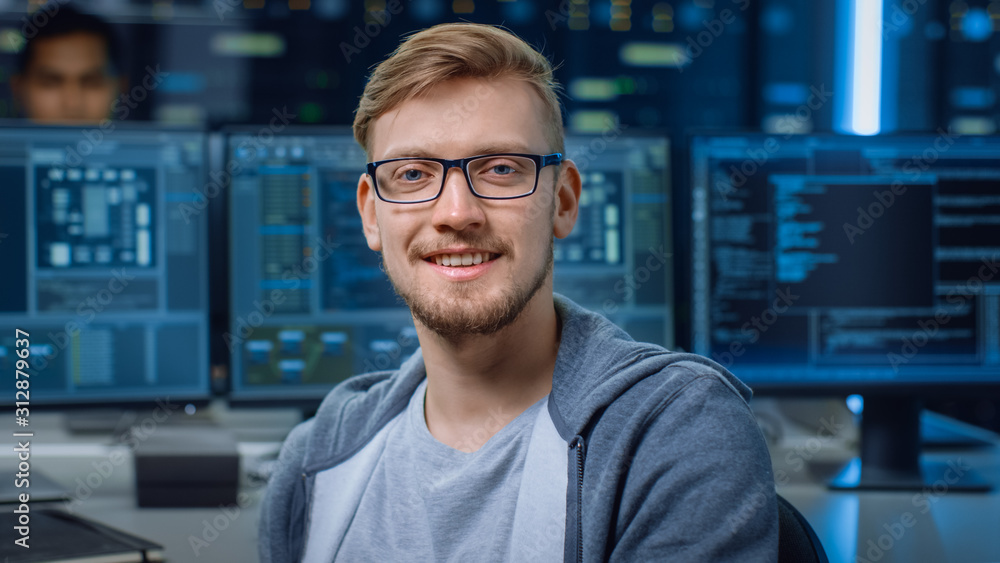 一位戴着眼镜微笑的聪明英俊的IT专家的肖像，在他身后的个人电脑