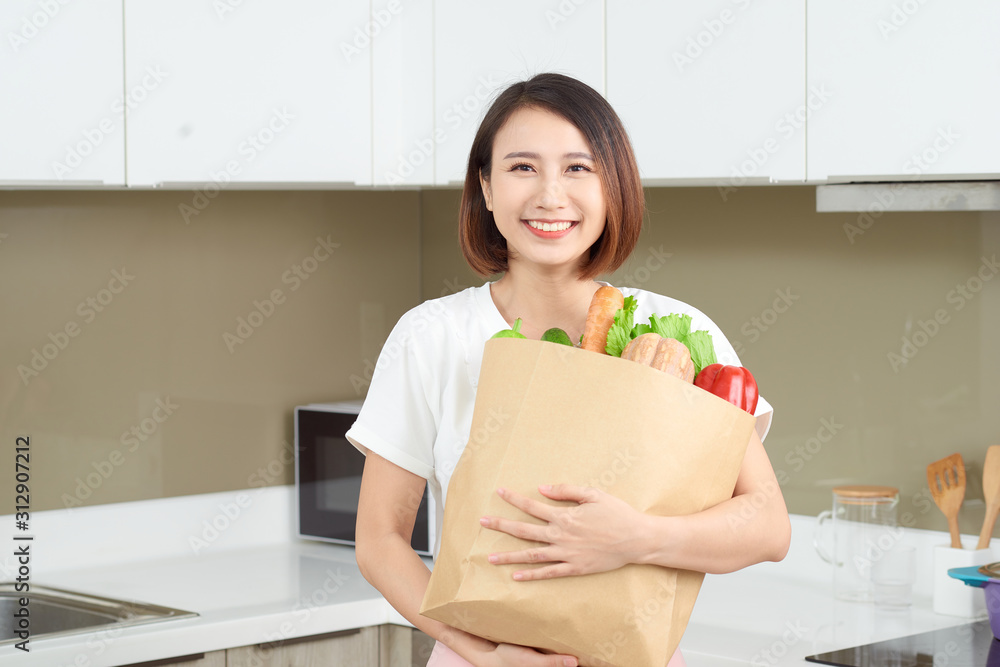 年轻的亚洲妇女在厨房里拿着装有蔬菜的杂货购物袋。