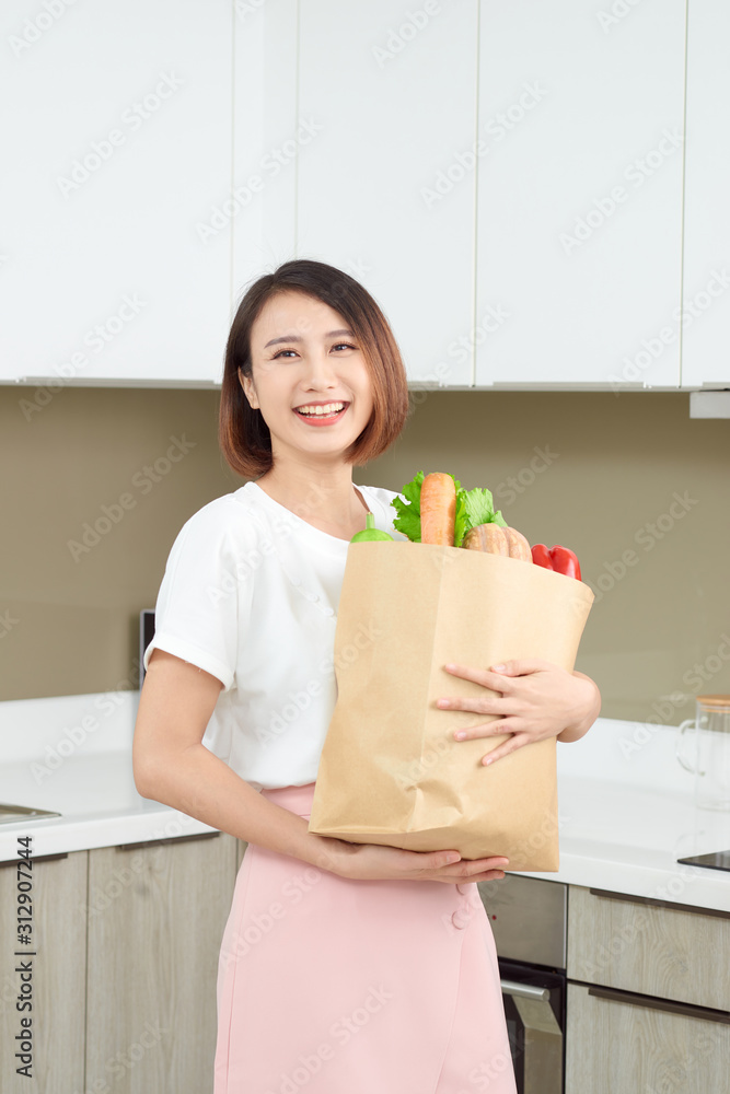 快乐、开朗的亚洲女人拿着装满新鲜产品、蔬菜、绿色蔬菜、法棍面包的纸袋