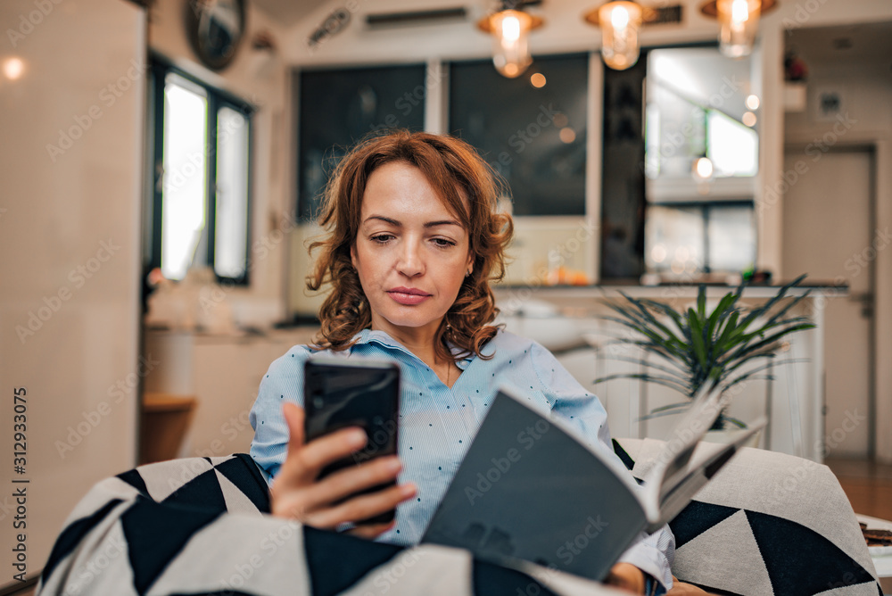 一位女士在家看书时用智能手机阅读短信的照片。