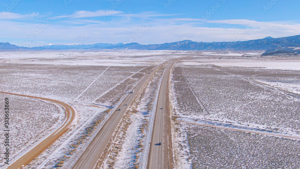 AERIAL: Flying along an asphalt highway leading vehicles across the snowy desert