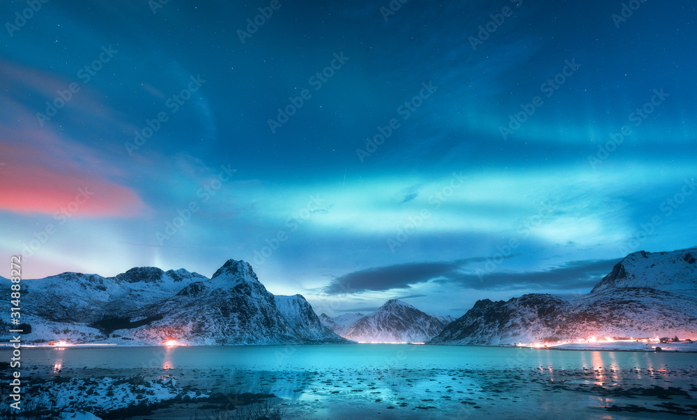 Aurora borealis over the sea coast, snowy mountains and city lights at night. Northern lights in Lof