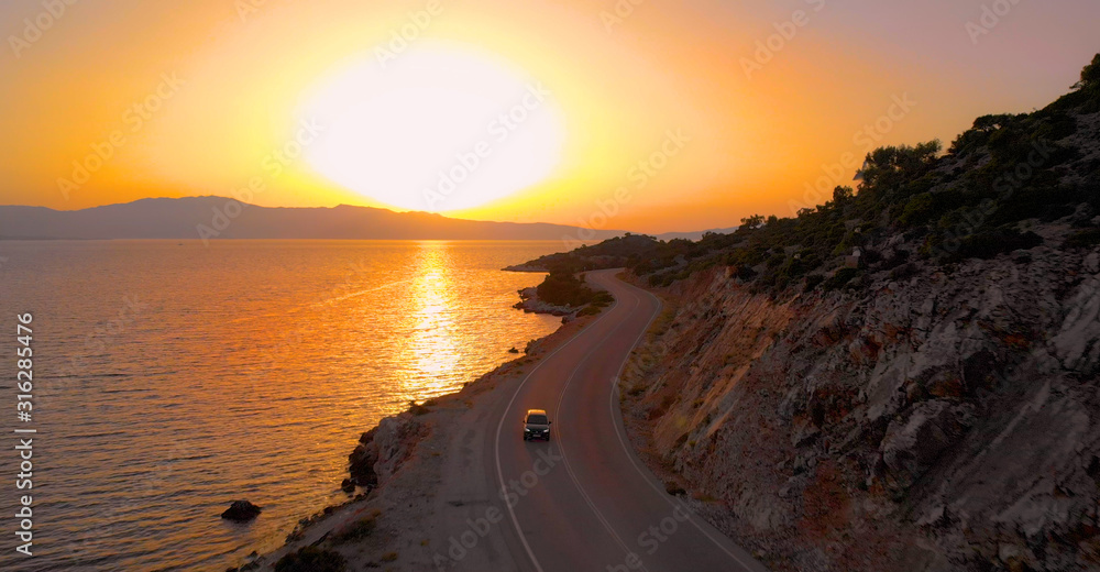 AERIAL: Dark colored car cruises along coastal road illuminated by the sunset.