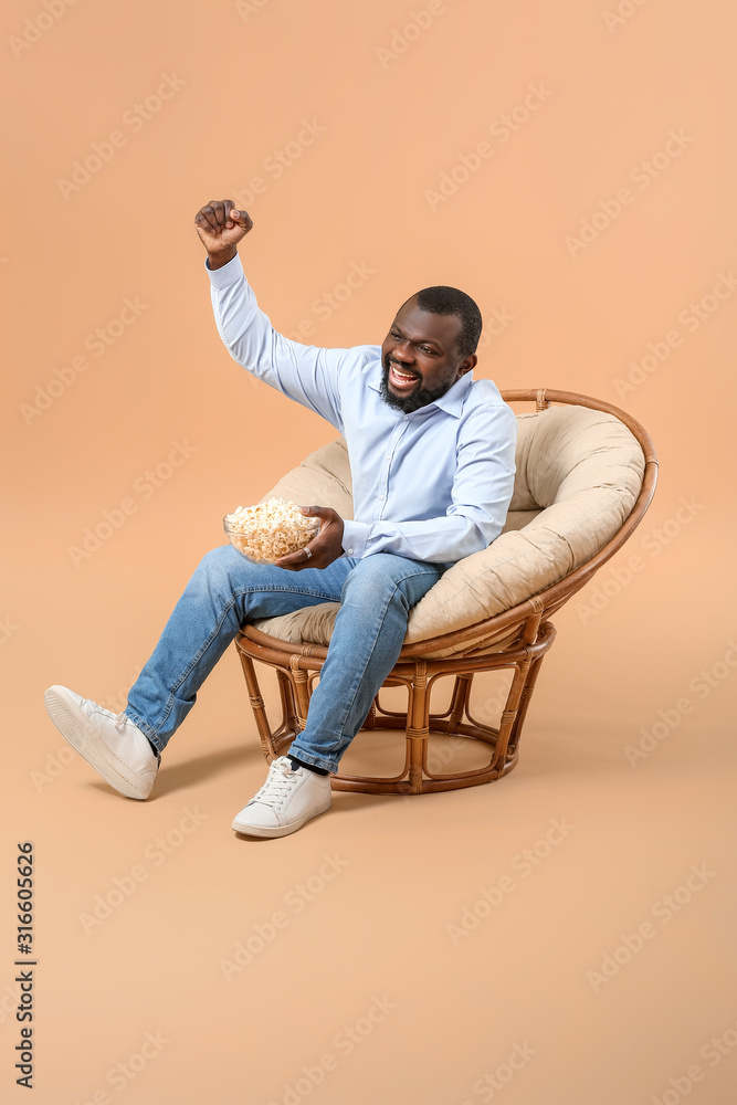 情绪激动的非裔美国男子拿着爆米花坐在扶手椅上看电视，背靠彩色靠背
