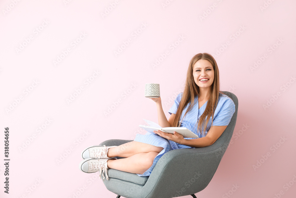 美丽的年轻女子坐在彩色墙上的扶手椅上看书喝茶