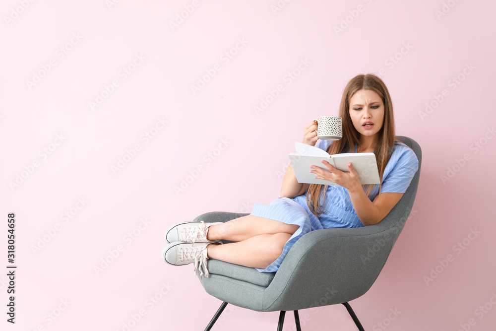 美丽的年轻女子坐在靠近彩色墙的扶手椅上看书喝茶