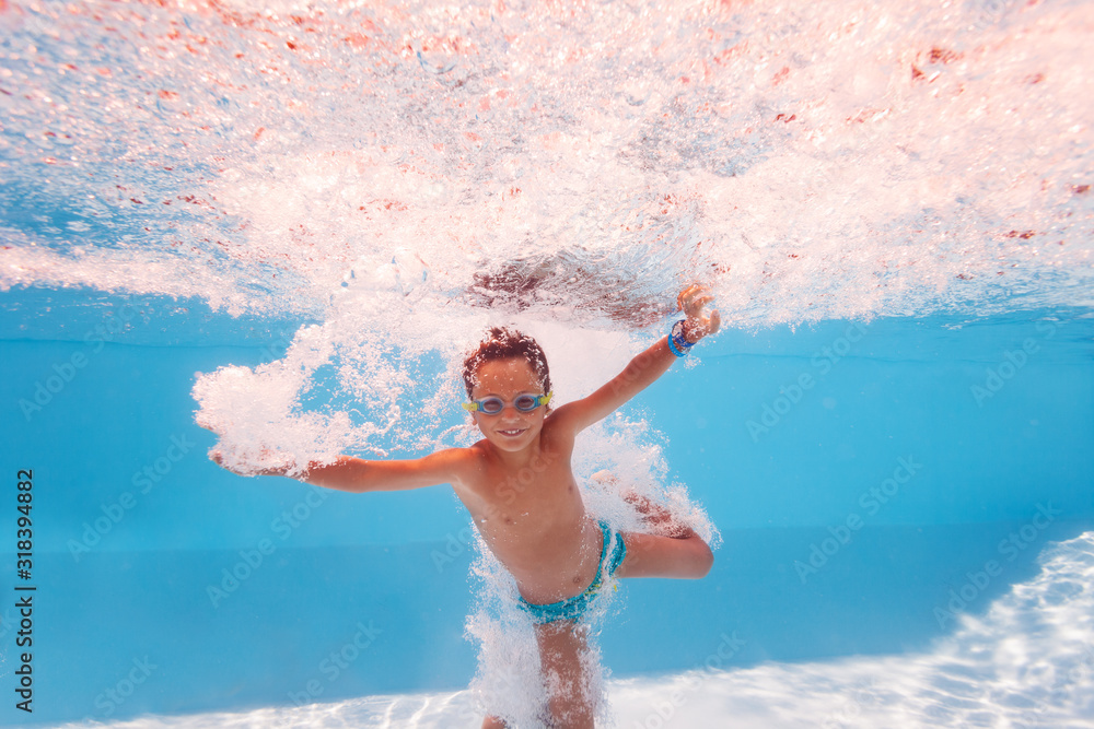 Happy little boy splash into the pool after jump making many bubbles, smiling and wearing swimming g