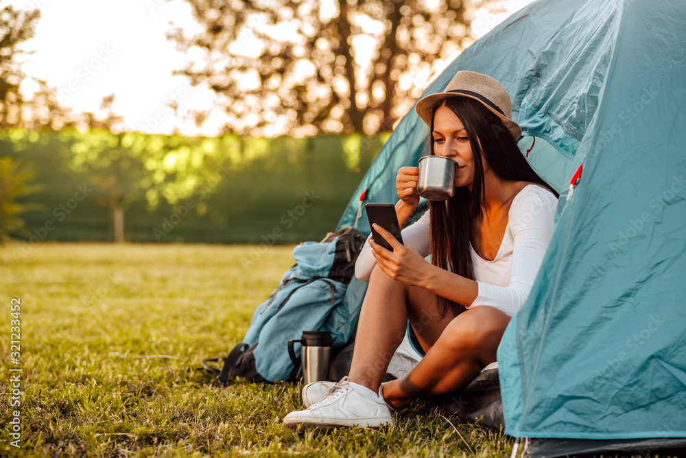 Portrait of a young woman at camping, sitting at tent drinking coffee and looking at smart phone.