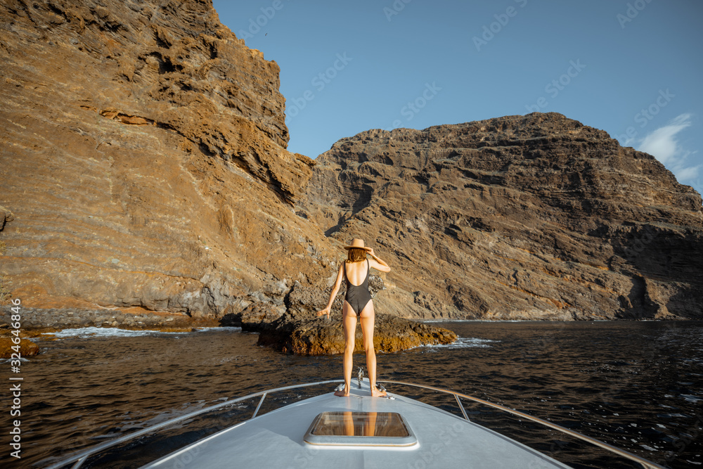 一名女子正在享受海上航行，站在游艇机头上，在令人惊叹的岩石海岸附近航行