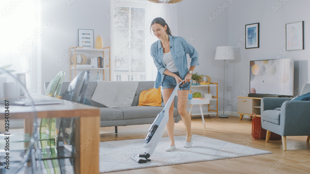 穿着牛仔裤、衬衫和短裤的年轻美女正在明亮舒适的房间里用吸尘器清洁地毯