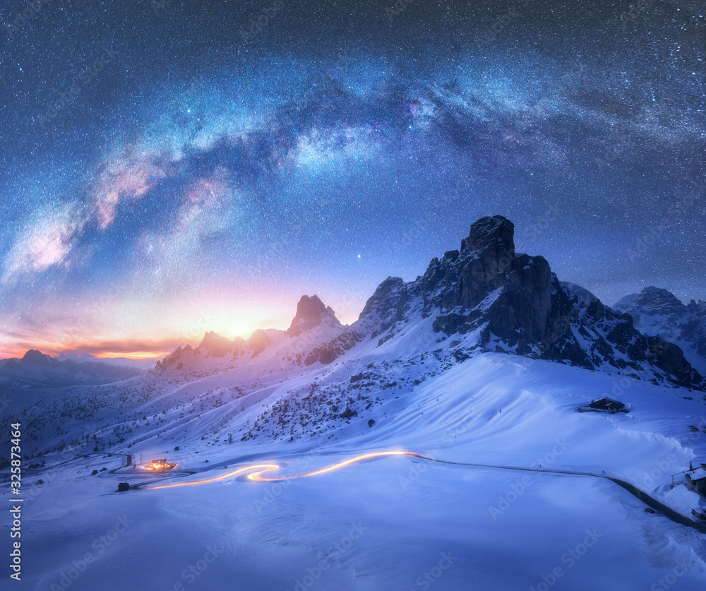 冬天夜晚，雪山上的银河和蜿蜒道路上模糊的汽车前灯。Be