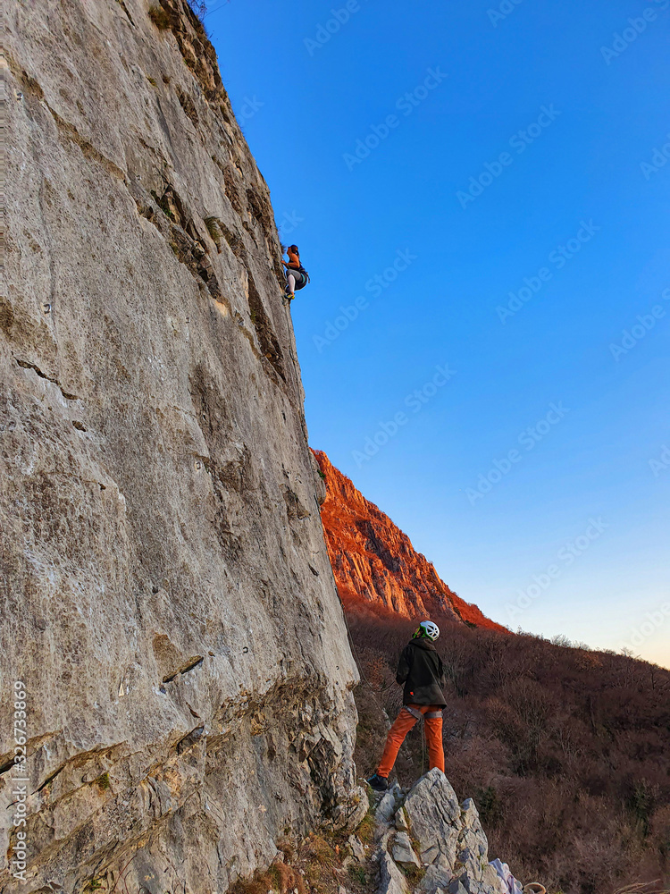 VERTICAL: Man looks up at female friend climbing up a rocky wall at sunset.