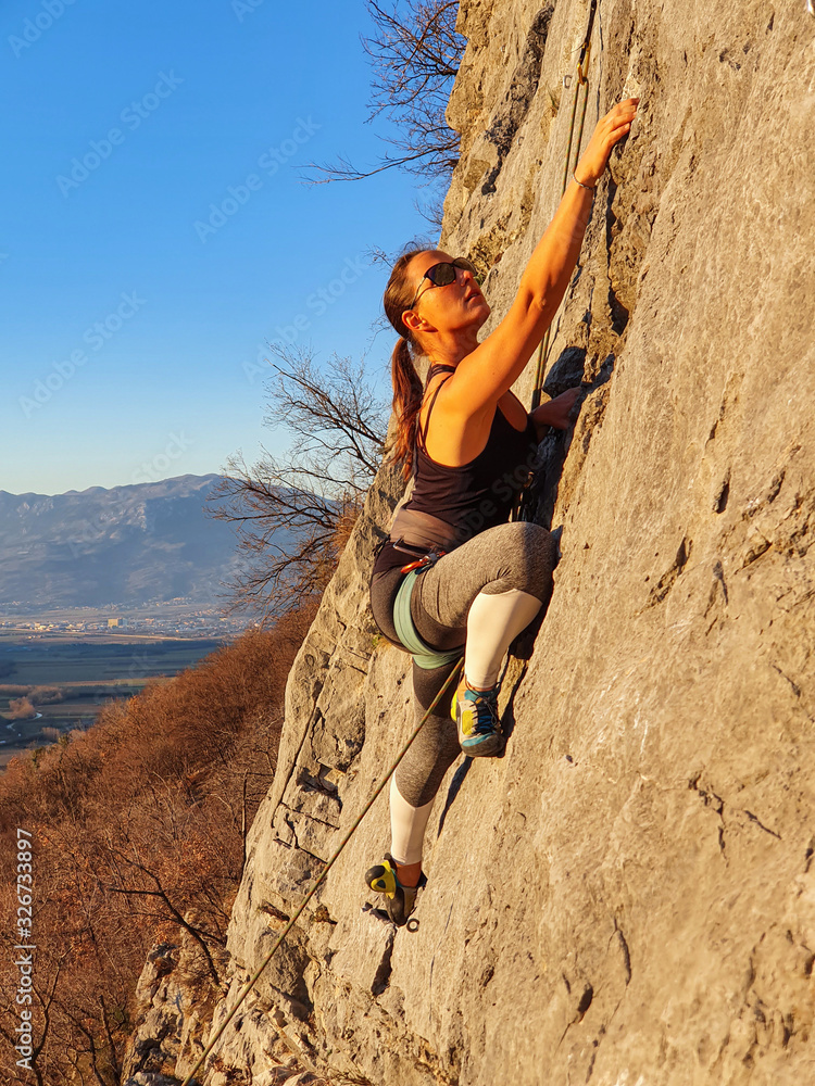 VERTICAL: Athletic woman wearing sunglasses climbs up a challenging rocky wall.