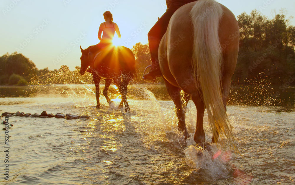 CLOSE UP: Two riders riding dark brown and palomino horse along river at sunset