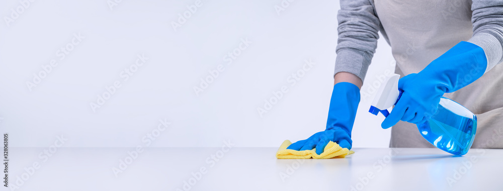 Young woman housekeeper is doing cleaning white table in apron with blue gloves, spray cleaner, wet 
