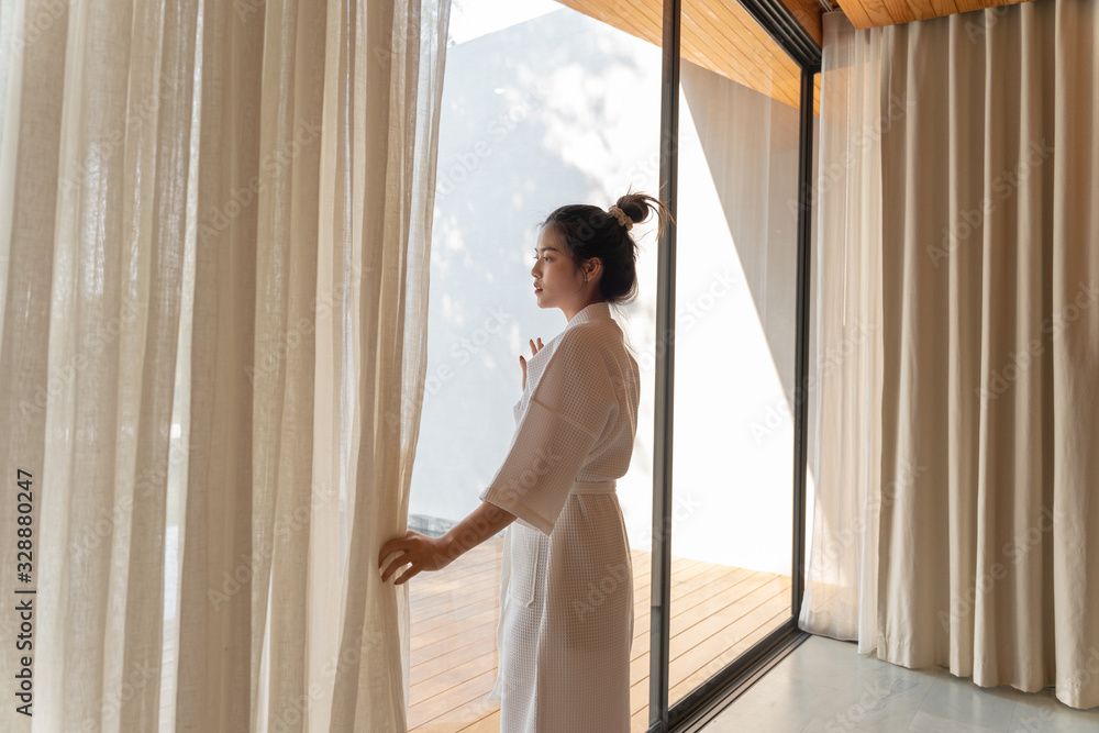 Asian woman in bathrobe suit open curtain to see view outside in morning, Travel and Holiday concept