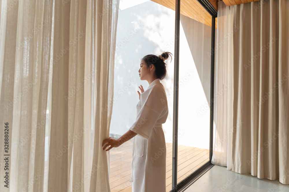 穿着浴袍套装的亚洲女性早上打开窗帘看外面的景色，旅行和度假概念