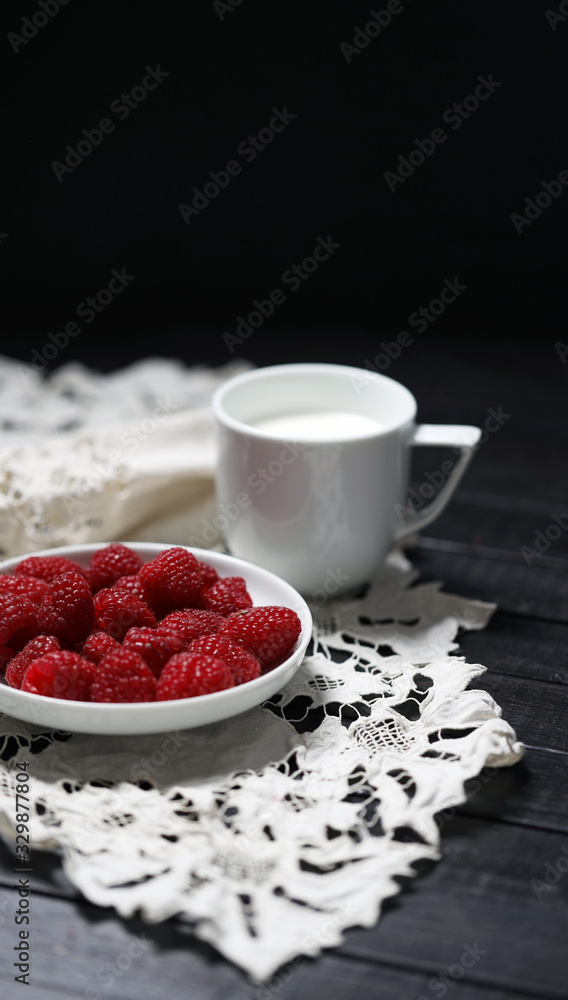 把成熟的树莓放在白色盘子里，把一杯牛奶放在放在木桌旁的白色手巾上