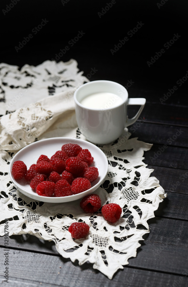 将成熟的覆盆子放在白色盘子里，三个浆果放在一边，在白色手巾上放一杯牛奶。