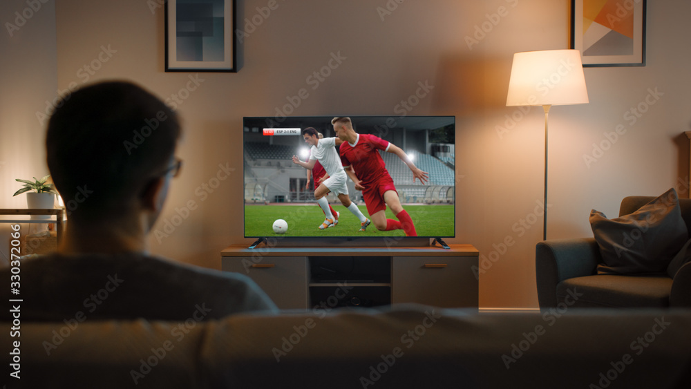 Young Man in Glasses is Sitting on a Sofa and Watching TV with a Soccer Match. Its Evening and Room