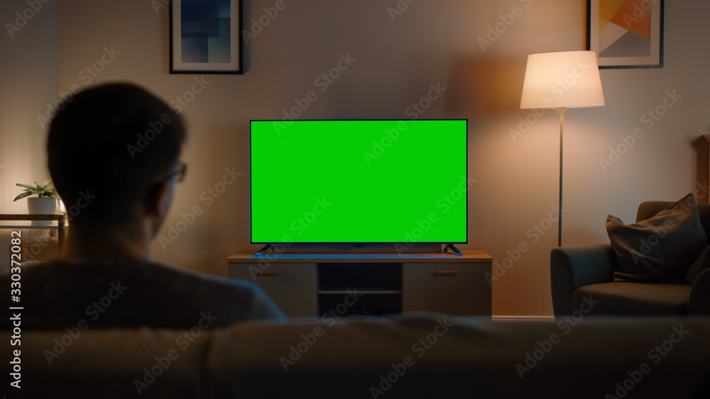 戴眼镜的年轻人坐在沙发上，用水平绿屏实物模型看电视。It